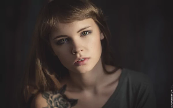 A close-up of a brunette woman with striking green eyes and a visible tattoo, showcasing her intense expression in soft lighting. This HD wallpaper features Anastasiya Scheglova.