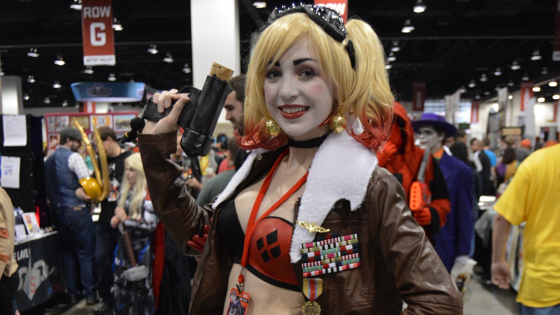 A woman in detailed cosplay with blonde pigtails and a brown jacket poses with a prop gun at a crowded convention, captured in 4K Ultra HD for PC desktop wallpaper.