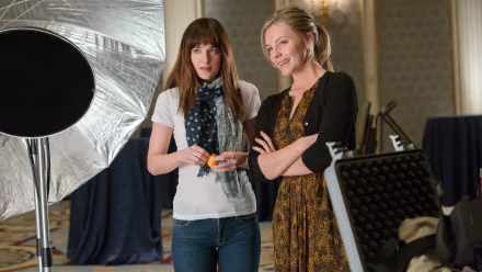 Dakota Johnson on the set of Fifty Shades of Grey, a movie scene captured as a HD desktop wallpaper and background. Two women engage in a conversation in a well-lit room with film equipment around.