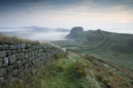 2K Quad HD desktop wallpaper: misty dawn over the man-made Hadrian's Wall winding across grassy hills, stone ramparts in foreground and ridgelines fading into fog.