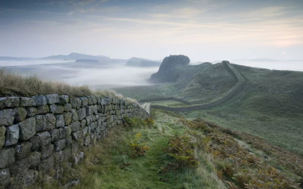 2K Quad HD desktop wallpaper: misty dawn over the man-made Hadrian's Wall winding across grassy hills, stone ramparts in foreground and ridgelines fading into fog.