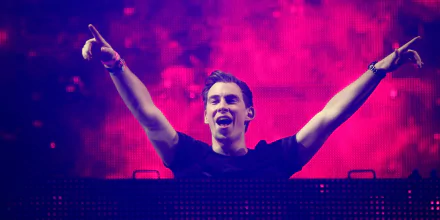 HD PC desktop wallpaper of a DJ on stage with arms raised, vivid pink-purple lights and a cheering crowd during a music performance.