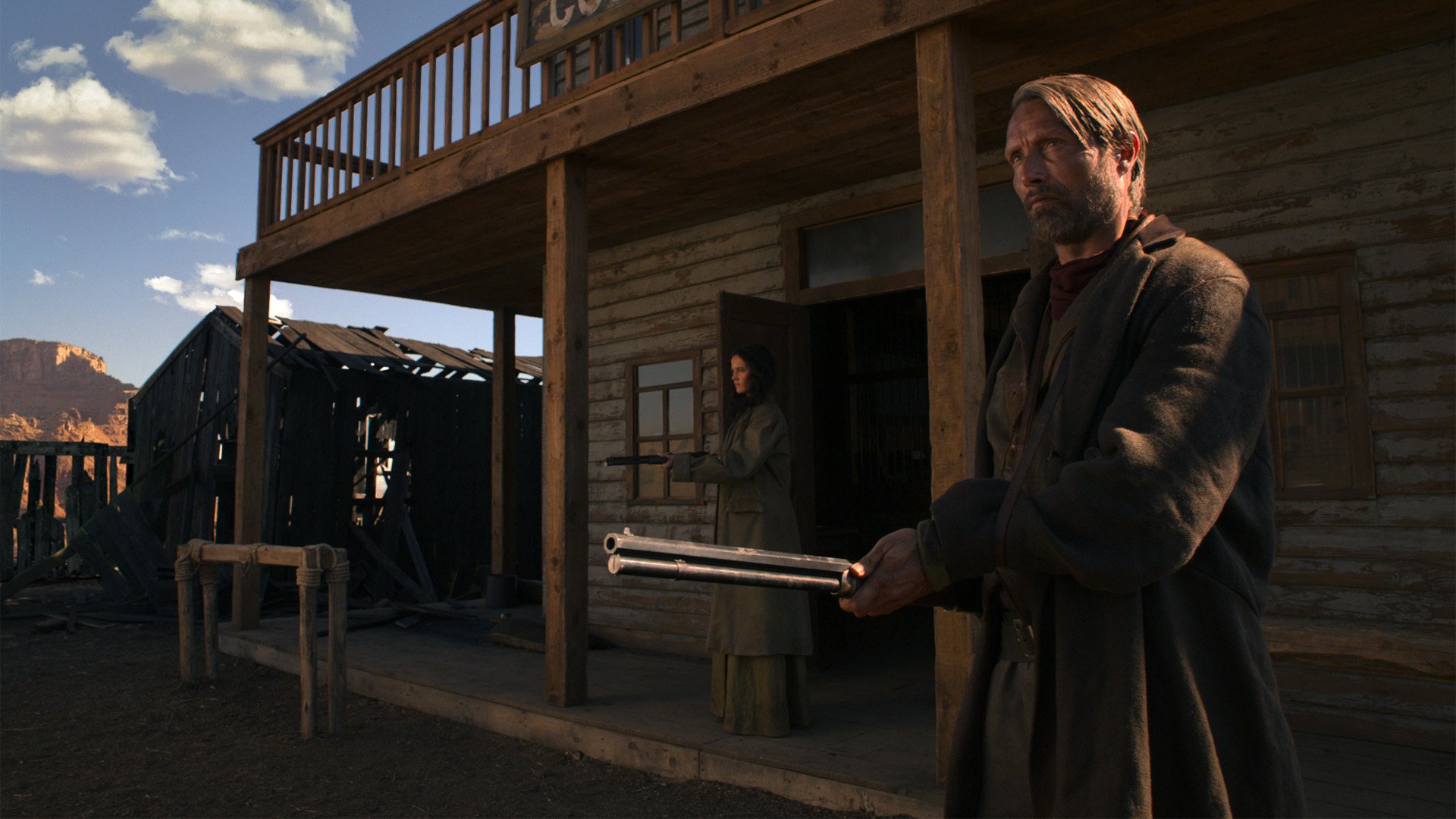 HD desktop wallpaper from the film The Salvation: a rugged man holds a double-barrel shotgun on a dusty western street while a woman watches from a saloon doorway.