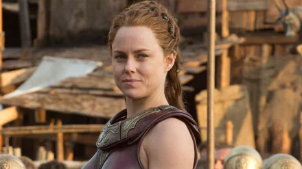 HD PC desktop wallpaper of a female warrior character in armor from the movie Hercules (2014), standing before a rustic wooden village with a determined expression.