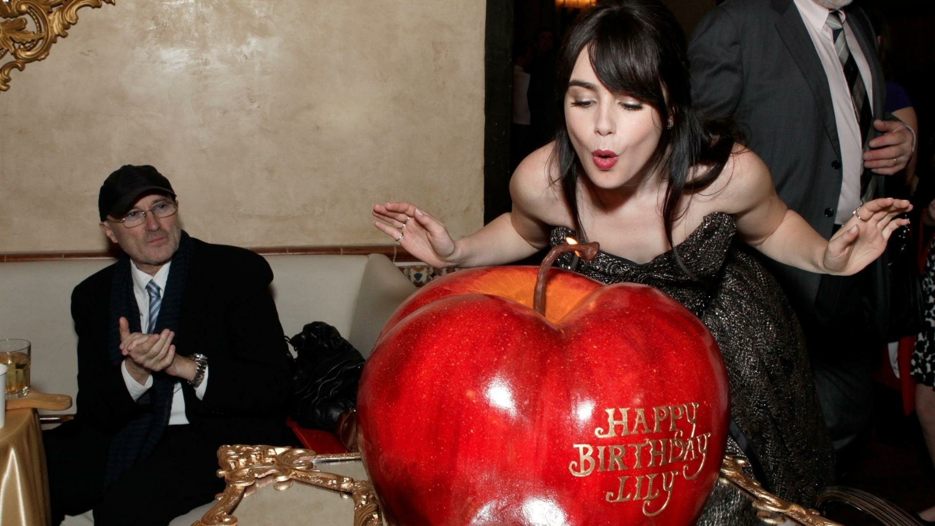 HD PC desktop wallpaper of a celebrity blowing out candles on a giant red apple birthday cake at a party.