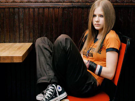 A stylish HD wallpaper featuring Avril Lavigne, sitting casually with her legs crossed, wearing an orange shirt and black pants against a wooden backdrop.