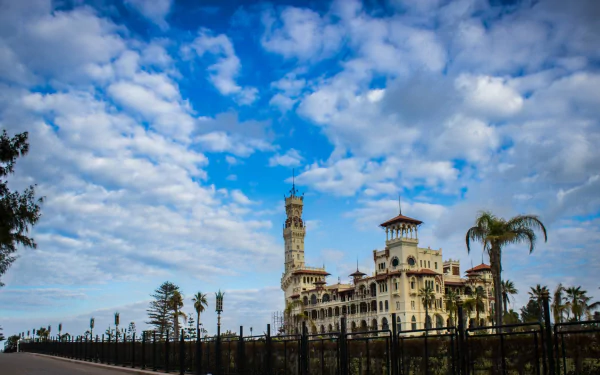 HD PC desktop wallpaper of Montaza Palace: a man-made ornate coastal palace with tower and palm trees beneath a bright, cloud-speckled blue sky.