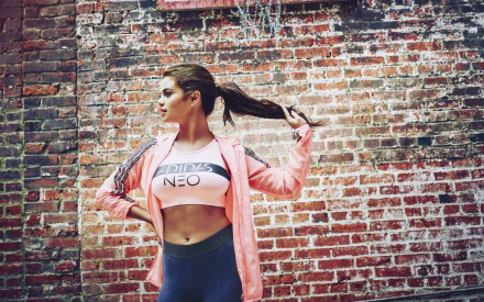 Selena Gomez in Adidas activewear stands confidently against a rustic brick wall, embodying a stylish blend of fitness and music culture. HD desktop wallpaper and background.