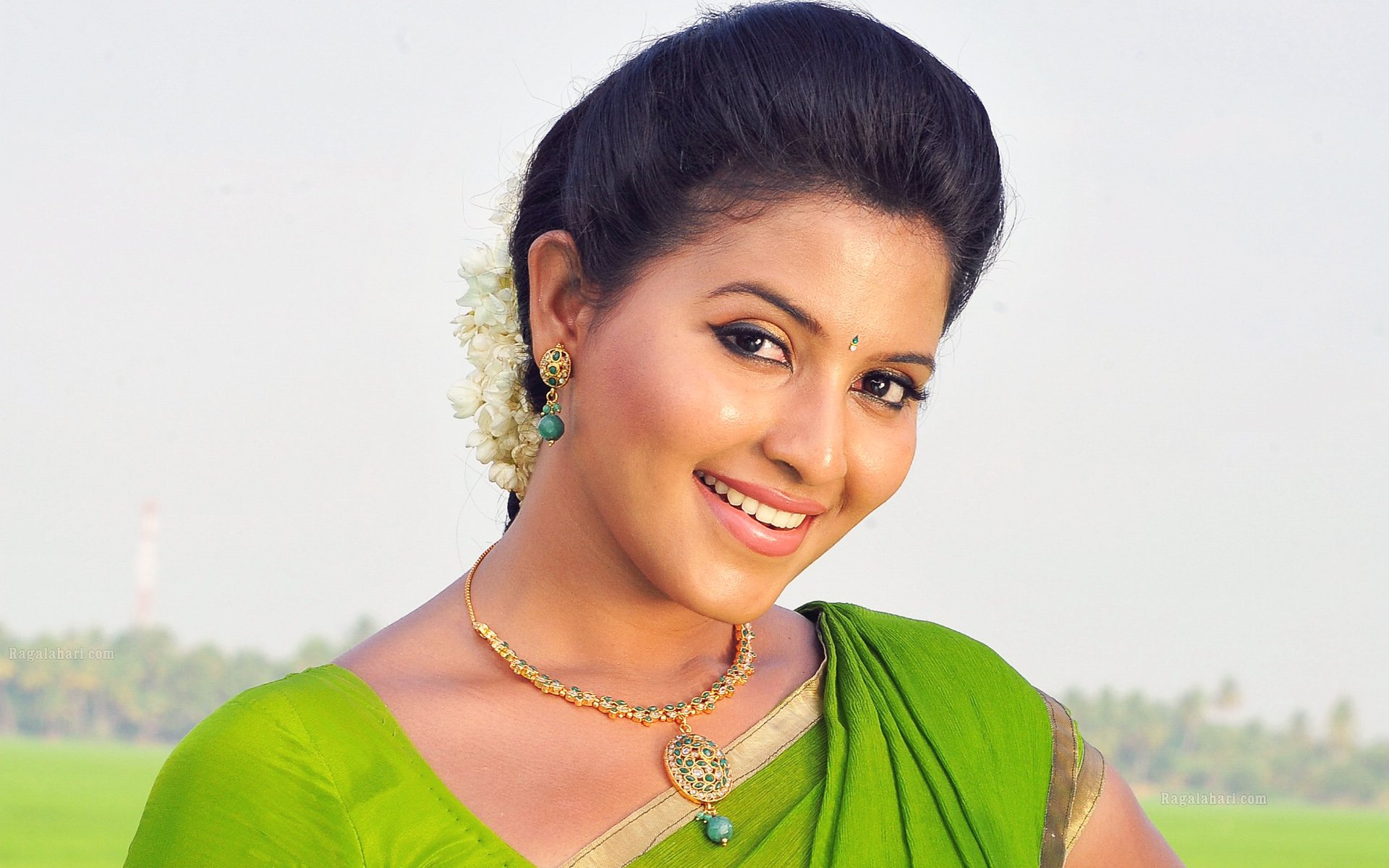 HD PC desktop wallpaper featuring a smiling woman named Anjali, dressed in a green saree with traditional jewelry against a soft outdoor background.