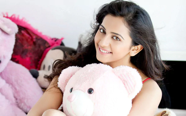 HD desktop wallpaper featuring celebrity Rakul Preet Singh smiling while holding a pink teddy bear amidst soft plush toys.