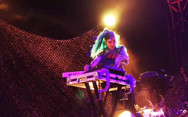 HD PC desktop wallpaper of musician Grimes performing live, illuminated by vibrant stage lighting and surrounded by electronic music equipment.