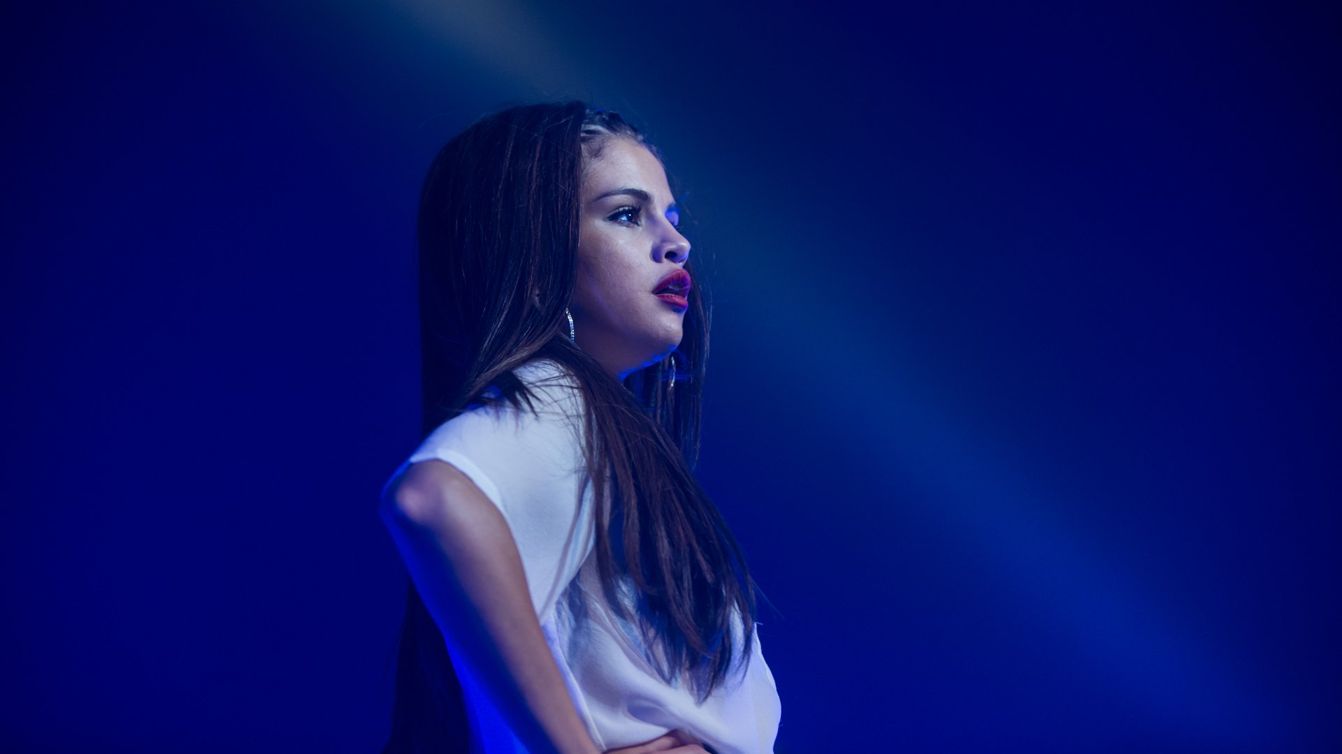 A striking HD wallpaper featuring Selena Gomez, captured in profile against a blue-lit background, embodying the essence of music and performance.