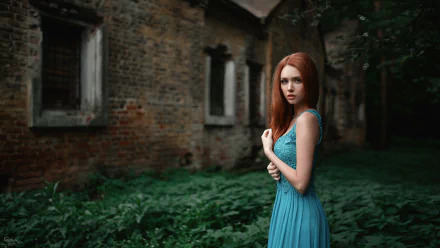 HD desktop wallpaper featuring a model with long red hair in a flowing blue dress, standing against a rustic brick building and lush greenery background.