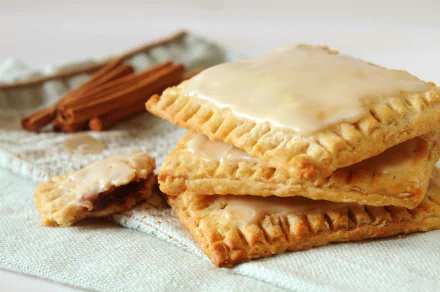 Close-up of glazed pop tarts stacked on a cloth with cinnamon sticks in the background, captured in 4K Ultra HD as a PC desktop wallpaper and background.