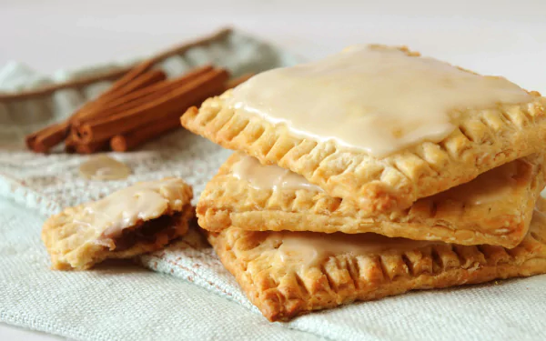 Close-up of glazed pop tarts stacked on a cloth with cinnamon sticks in the background, captured in 4K Ultra HD as a PC desktop wallpaper and background.