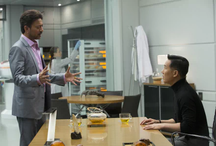 A scene from Jurassic World featuring B.D. Wong and Irrfan Khan engaged in a serious conversation at a modern table, with drinks and scientific equipment in the background.