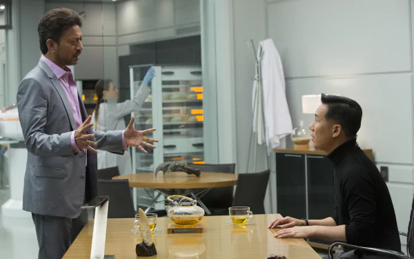 A scene from Jurassic World featuring B.D. Wong and Irrfan Khan engaged in a serious conversation at a modern table, with drinks and scientific equipment in the background.
