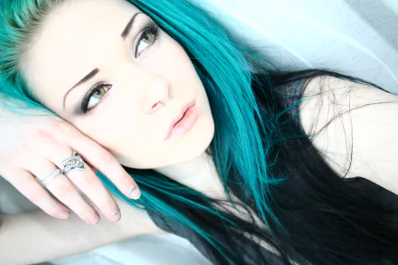 A woman with striking blue hair gazes thoughtfully, resting her head on her hand. This HD wallpaper captures her expressive face in a serene setting.