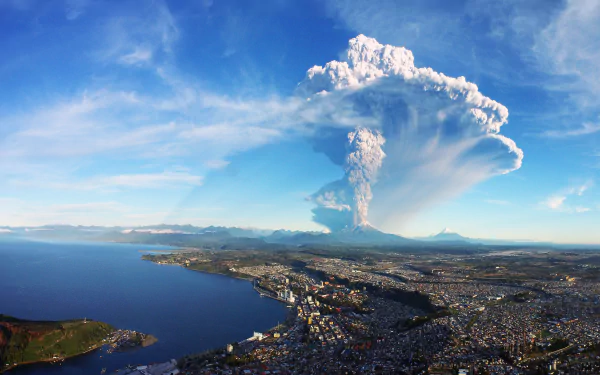 HD desktop wallpaper featuring a stunning view of a volcanic eruption with a city, lake, and mountains in the foreground under a clear blue sky. The eruption creates a dramatic plume of smoke and ash.