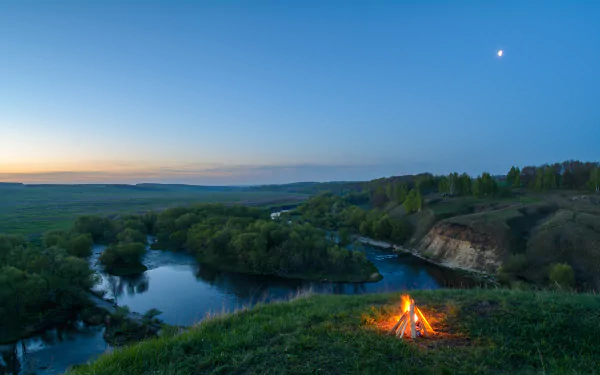 A serene landscape featuring a vibrant bonfire beside a tranquil river, surrounded by lush greenery under a twilight sky, perfect for a 4K Ultra HD desktop wallpaper.