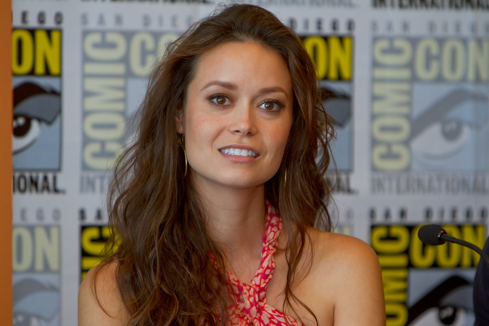 HD PC wallpaper: smiling female celebrity with long brown hair in a red patterned top at a Comic-Con panel, facing camera against a branded backdrop.