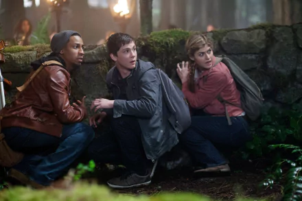 A dramatic scene from Percy Jackson: Sea of Monsters, featuring Alexandra Daddario, Logan Lerman, and Brandon T. Jackson crouched behind a stone wall in a mysterious forest setting.