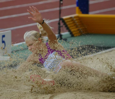 Track-and-field athlete landing in the long jump sandpit, sand flying around her — HD PC desktop sports wallpaper/background.