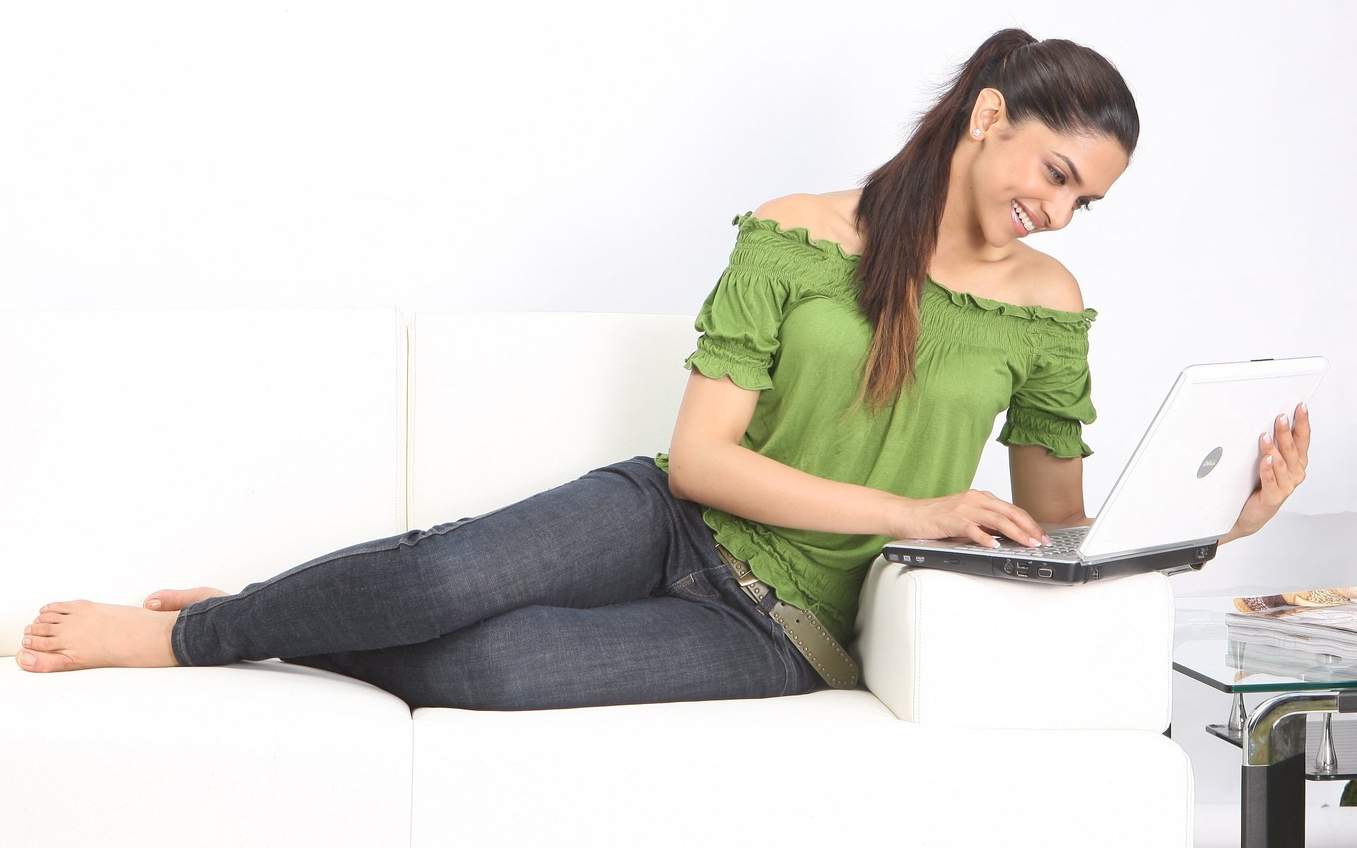 Deepika Padukone sits comfortably on a white sofa, wearing a vibrant green top and jeans, as she types on a laptop, showcasing a relaxed and cheerful atmosphere.