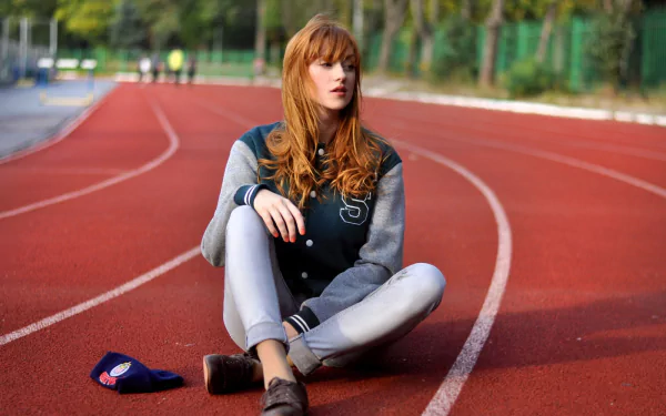 HD PC desktop wallpaper: red-haired woman sitting cross-legged on a running track, letterman jacket and cap beside her, trees and lanes receding behind.