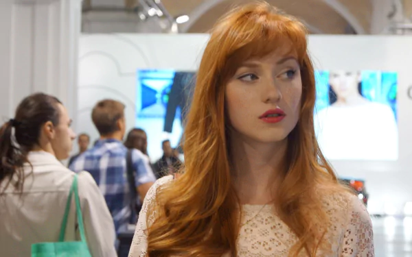 HD PC desktop wallpaper featuring a woman with long red hair, identified as Alina Kovalenko, standing indoors with people and bright screens in the background.