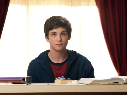 Logan Lerman sits at a desk with books in front of him, gazing thoughtfully. This HD wallpaper captures a moment from the movie The Perks of Being a Wallflower.