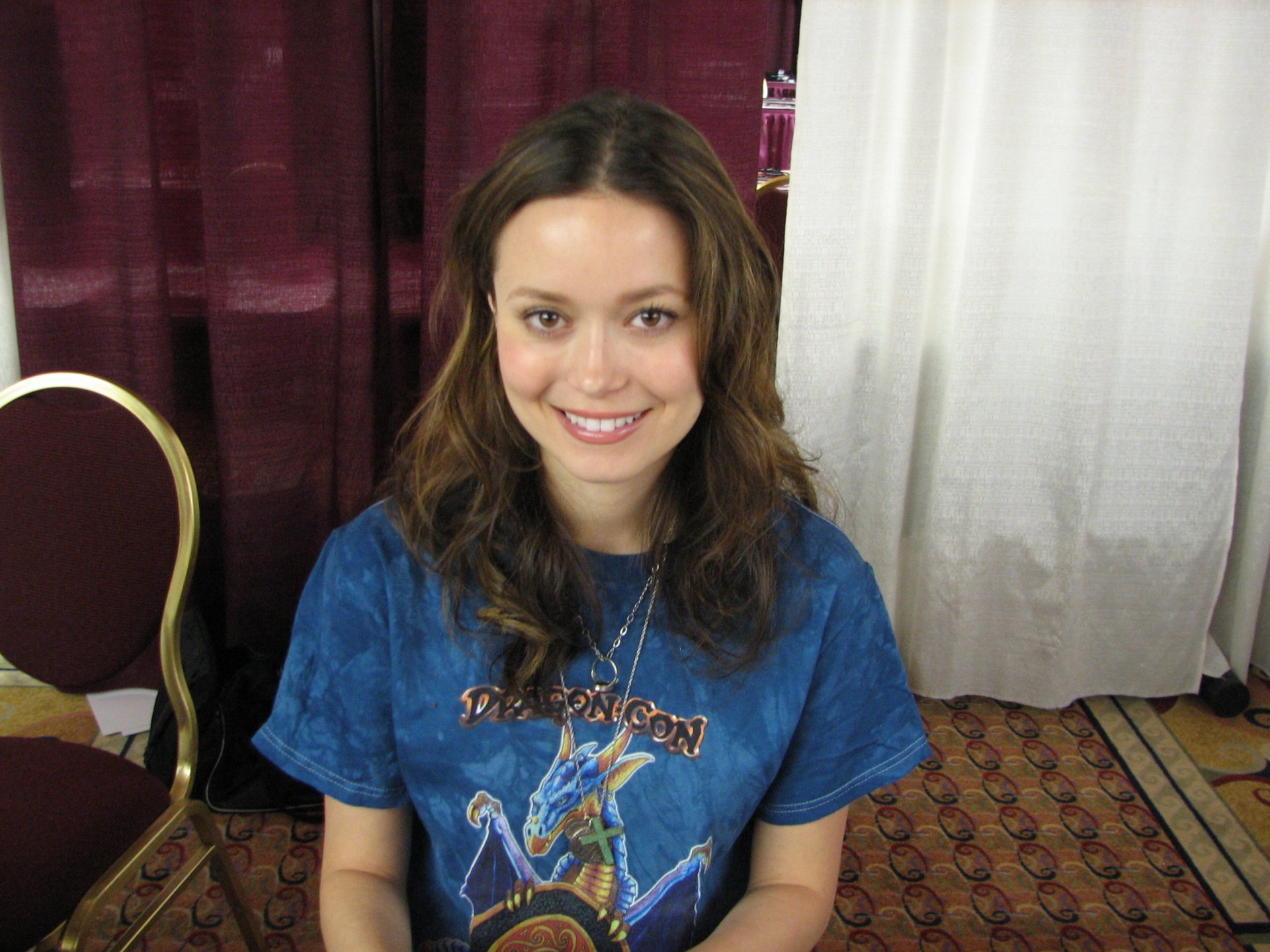 HD PC desktop wallpaper: portrait of a smiling young woman seated against curtains, wearing a blue graphic T-shirt and brown wavy hair.