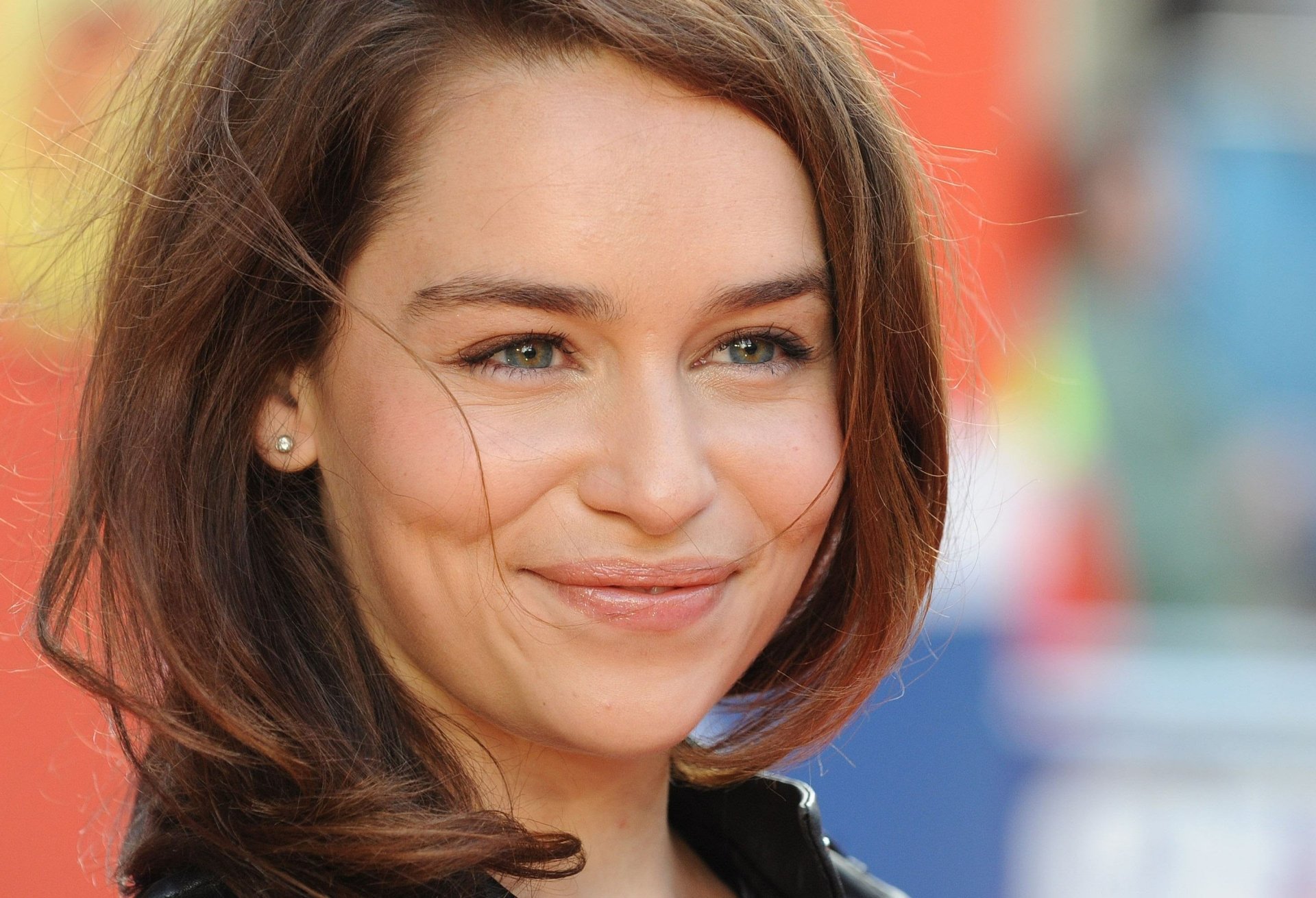 A close-up of Emilia Clarke smiling, showcasing her expressive eyes and wavy hair, set against a colorful background. This image serves as a vibrant HD desktop wallpaper.