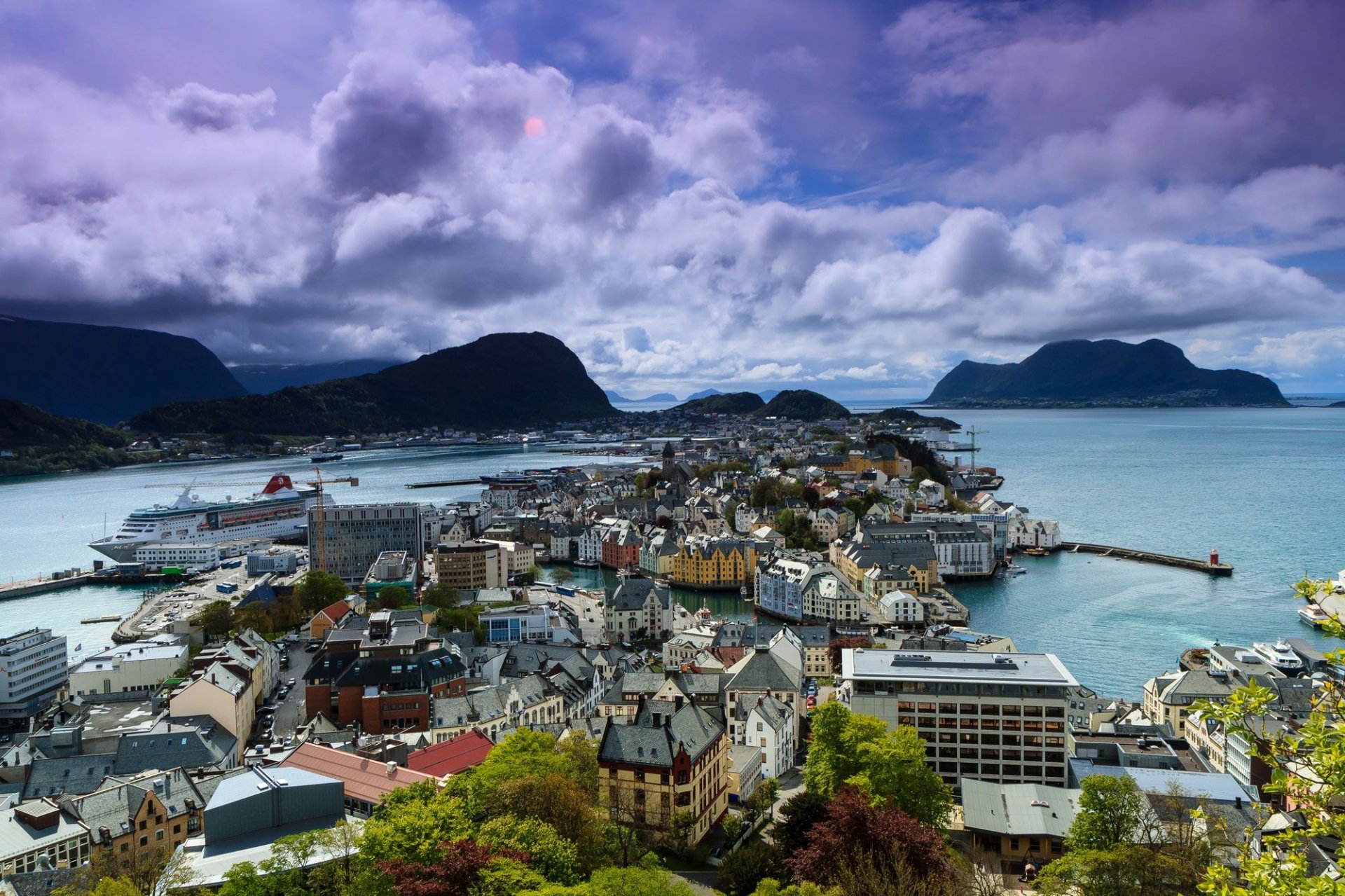 Aerial view of Ålesund, Norway, showcasing the town's man-made structures nestled between fjords and mountains under a dramatic cloudy sky.