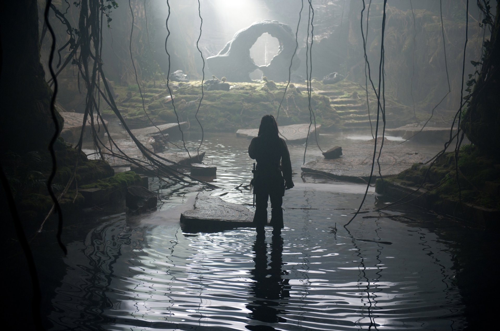 A 4K Ultra HD scene from Pirates of the Caribbean: On Stranger Tides showing a silhouetted figure standing in shallow water inside a cavern with hanging vines and a rock archway.