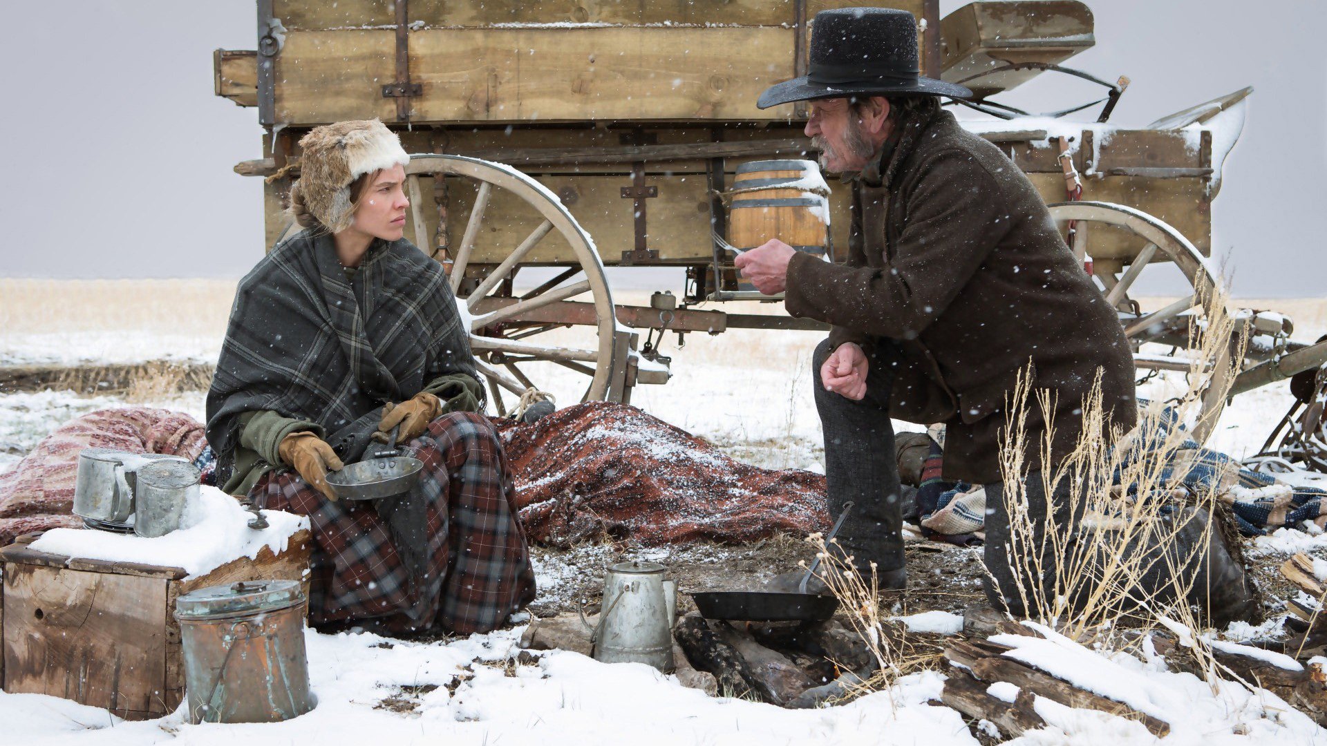 HD PC desktop wallpaper featuring Hilary Swank and Tommy Lee Jones in a snowy scene from the movie The Homesman, with period clothing and a wooden wagon in the background.