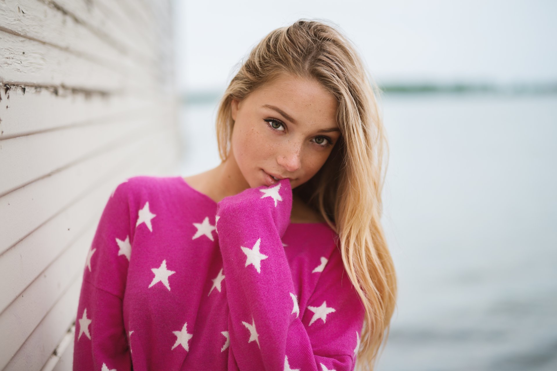 Blonde woman with green eyes and freckles wears a pink star-patterned sweater, posing thoughtfully against a blurred outdoor background in this 4K Ultra HD wallpaper.
