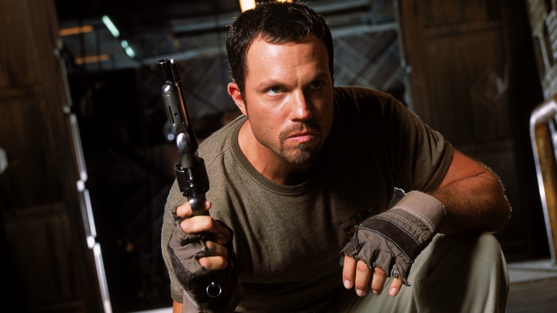 Man with a stern expression clutching a pistol in a dim industrial setting — moody action shot rendered as a 2K Quad HD PC desktop wallpaper.