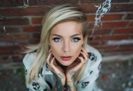A blonde woman with blue eyes and tattoos poses against a rustic brick wall, showcasing her unique style in this vibrant 4K Ultra HD wallpaper.