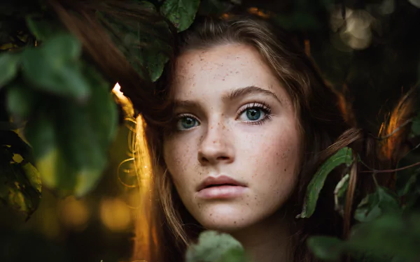 Close-up of a brunette model with blue eyes and freckles framed by leaves, captured in stunning 8K Ultra HD quality for a striking PC desktop wallpaper.