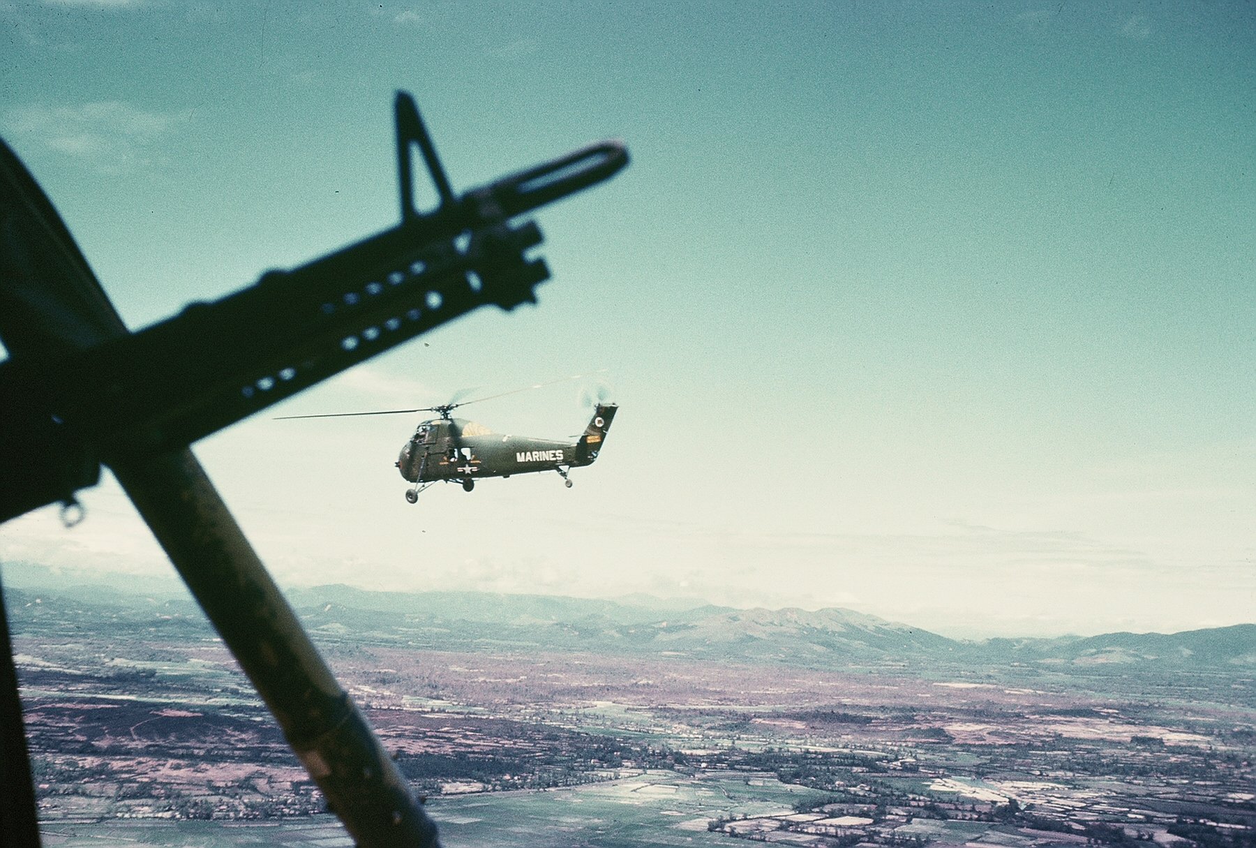 Military Sikorsky H-34 helicopter in flight over snowy terrain, framed from a cockpit with a gun silhouette — HD PC desktop wallpaper and background.
