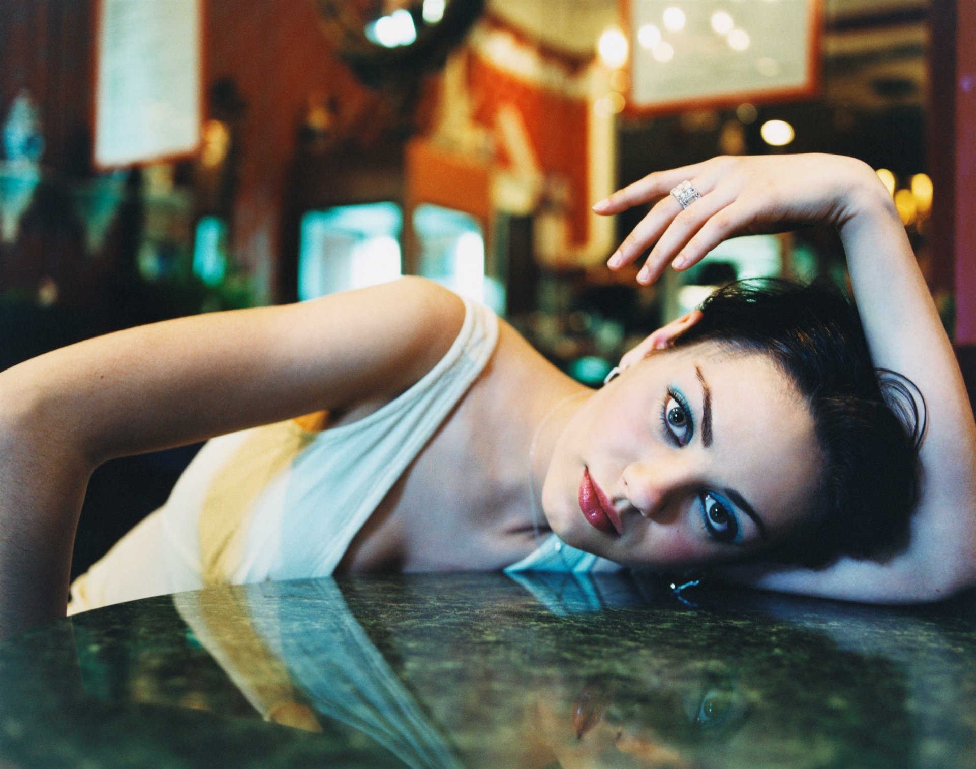 A captivating HD wallpaper featuring Mila Kunis, elegantly posed with her head resting on a table, exuding a striking gaze and a relaxed charm in a vibrant setting.