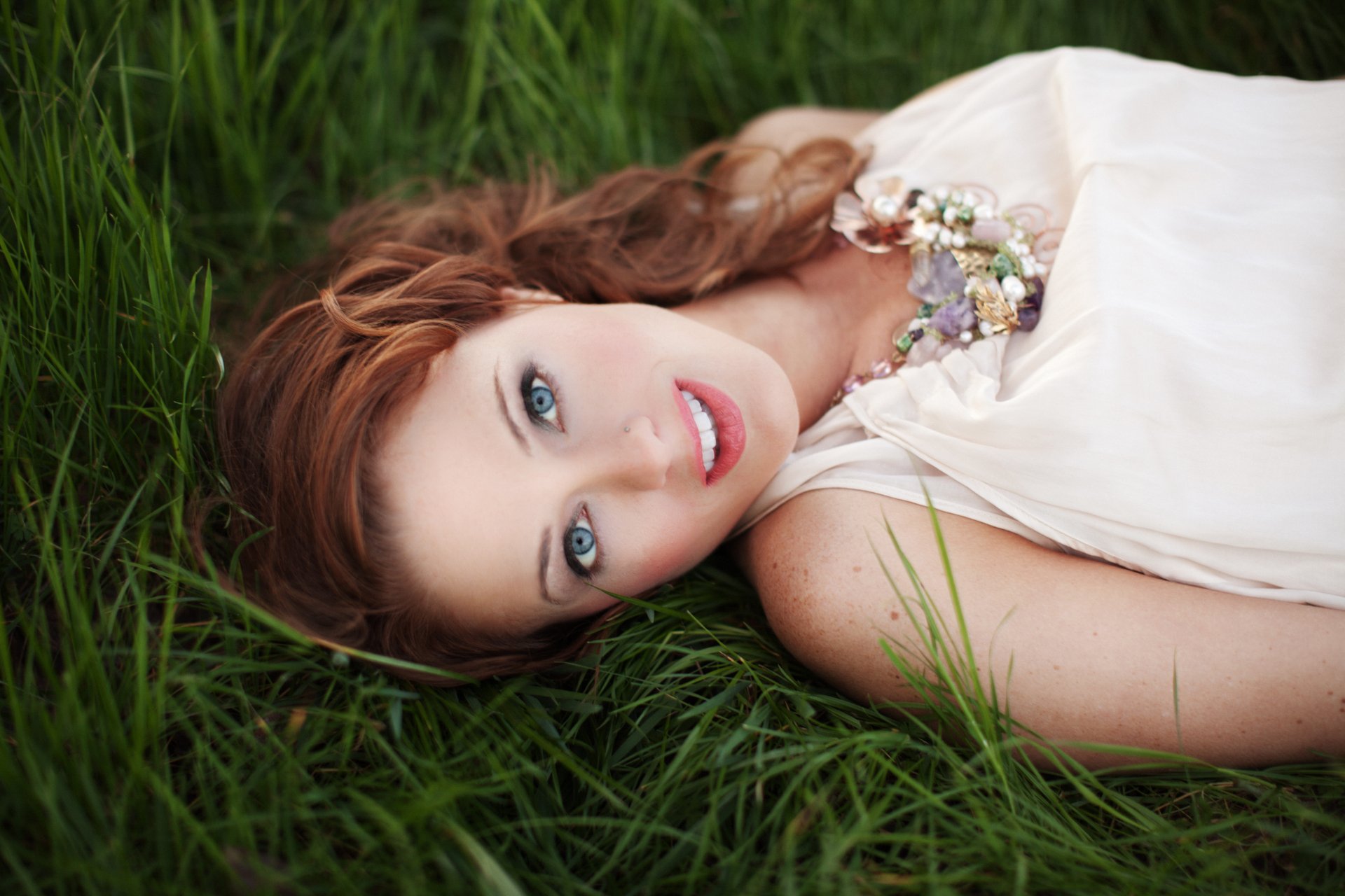 A woman model lies in lush green grass, showcasing her vibrant blue eyes and elegant attire. This stunning image is a 4K Ultra HD wallpaper and background.