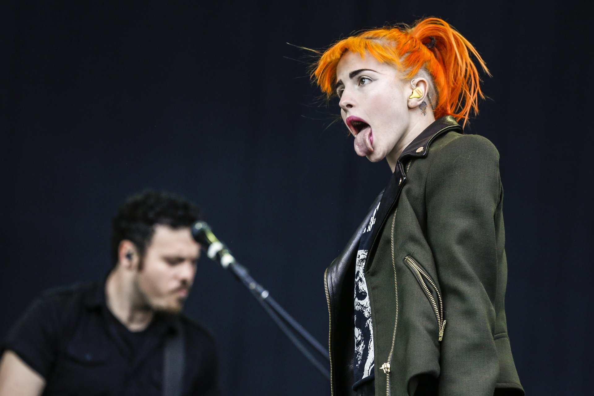 Female rock singer with bright orange hair mid-performance, tongue out, guitarist blurred behind; music-themed 2K Quad HD PC desktop wallpaper and background.