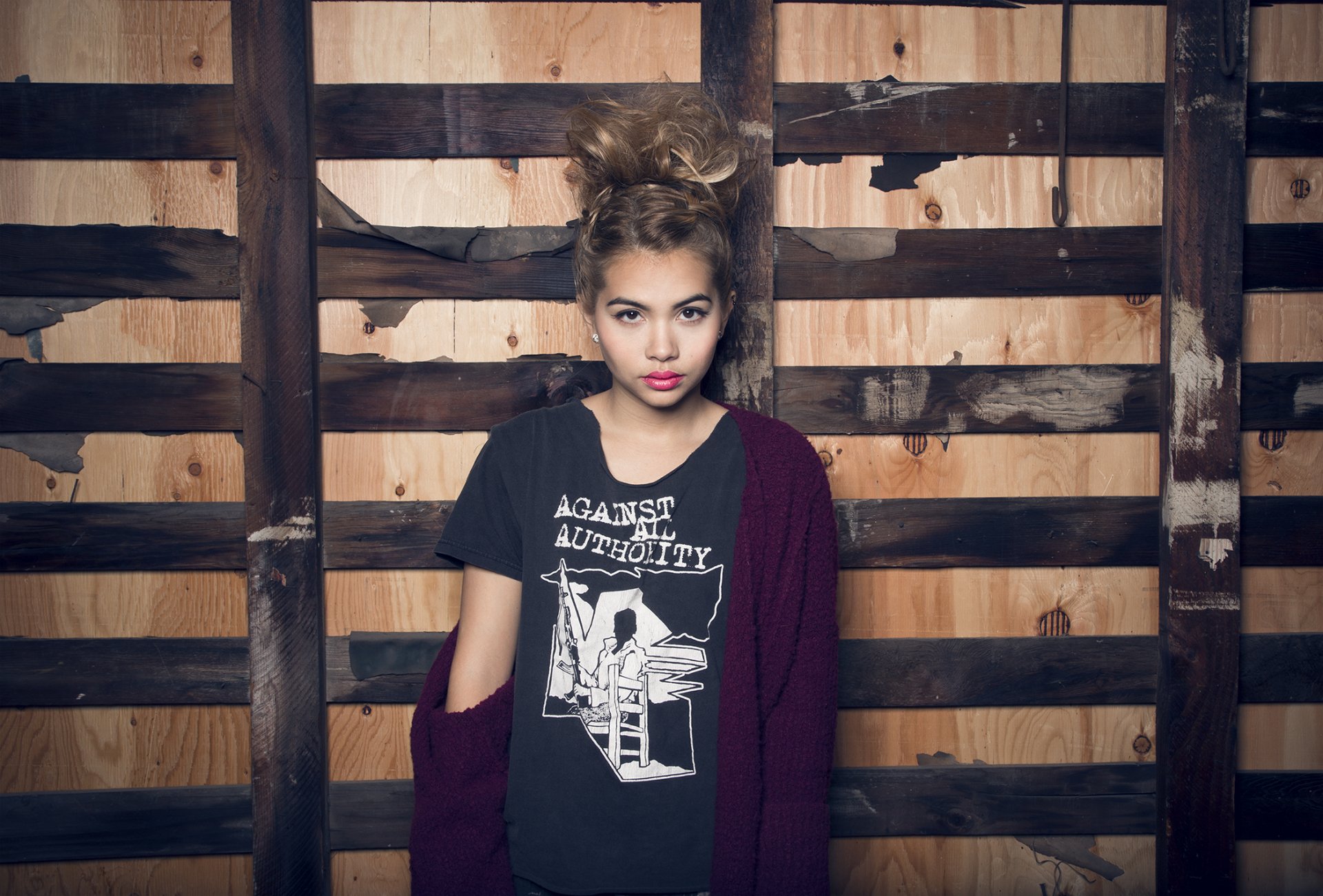 HD PC desktop wallpaper featuring celebrity Hayley Kiyoko standing against a rustic wooden wall, wearing a graphic tee and a deep red cardigan.