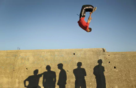 Parkour athlete mid-flip above a concrete wall with onlookers' shadows cast below — Sports action captured in a 4K Ultra HD PC desktop wallpaper/background.