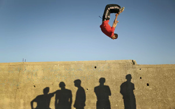 Parkour athlete mid-flip above a concrete wall with onlookers' shadows cast below — Sports action captured in a 4K Ultra HD PC desktop wallpaper/background.