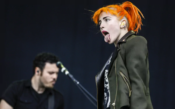 Female rock singer with bright orange hair mid-performance, tongue out, guitarist blurred behind; music-themed 2K Quad HD PC desktop wallpaper and background.