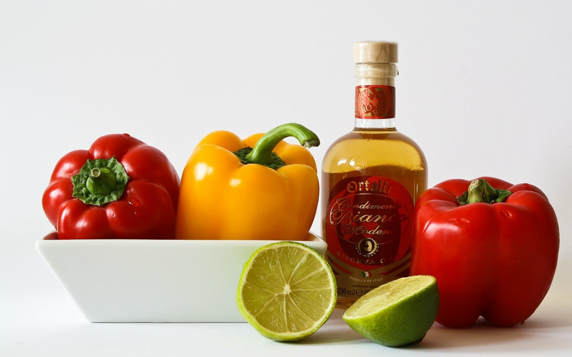 HD desktop wallpaper featuring a still life of vibrant red and yellow capsicums, a sliced lime, and a bottle of pepper sauce against a clean white background.