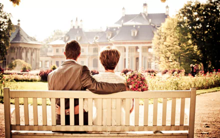A couple sits close together on a bench in a sunlit garden, overlooking a grand historic building, captured in an HD desktop wallpaper celebrating love and serene moments.